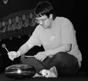 Person seated and playing a steel tongue drum with mallets in both hands on a dark stage background.